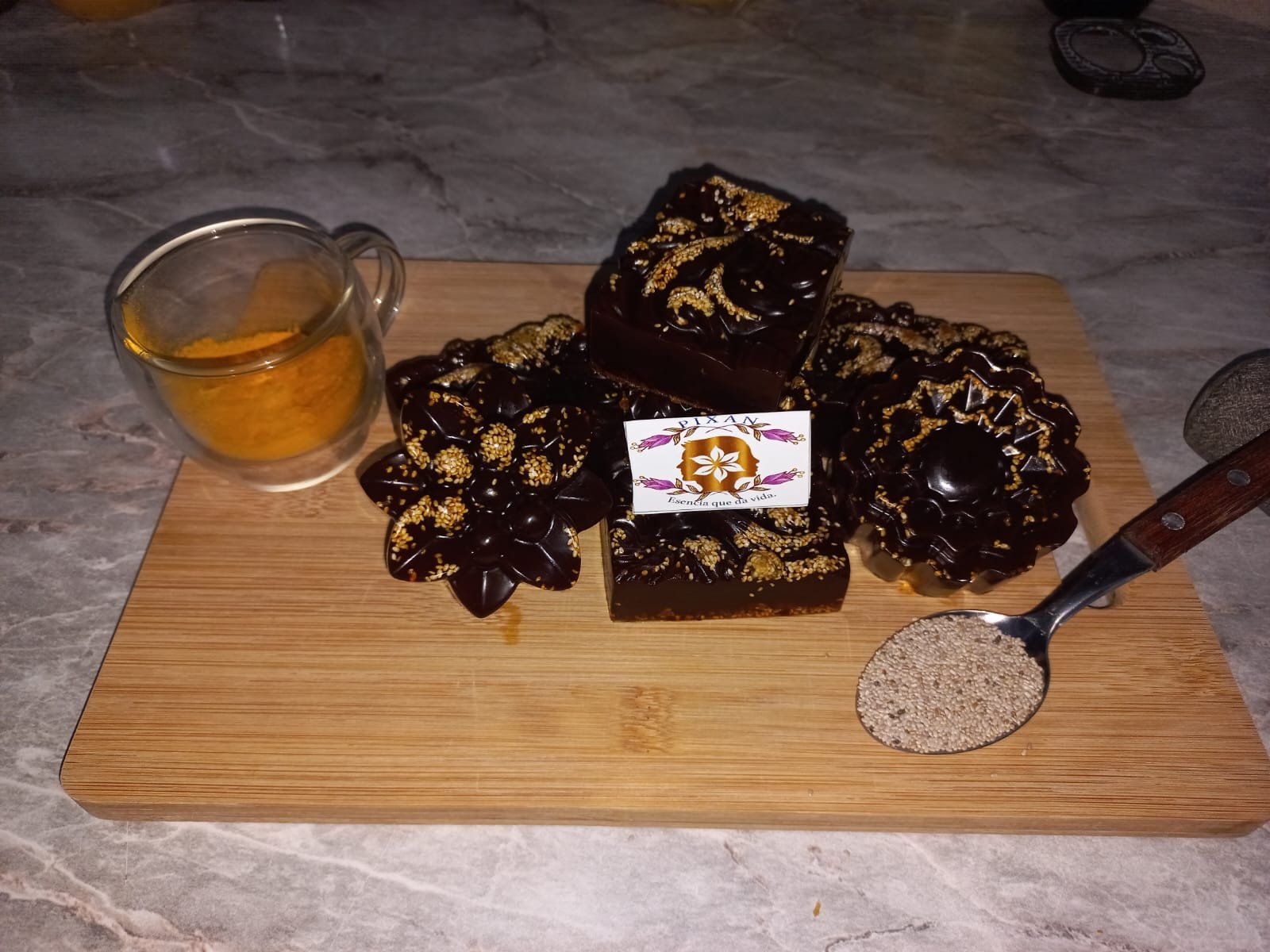 jabón pixani de curcuma con chia sobre tabla de madera en una barra de cocina junto a una taza con curcuma en polvo y una cuchara con chia.